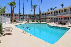 Una piscina con sillones de descanso y sombrillas, rodeada de palmeras y un edificio de varios pisos con balcones.