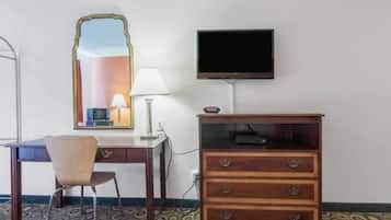 Ein Hotelzimmer mit Schreibtisch, Stuhl, Kommode, Spiegel, Lampe, Fernseher und einem gemusterten Teppich.