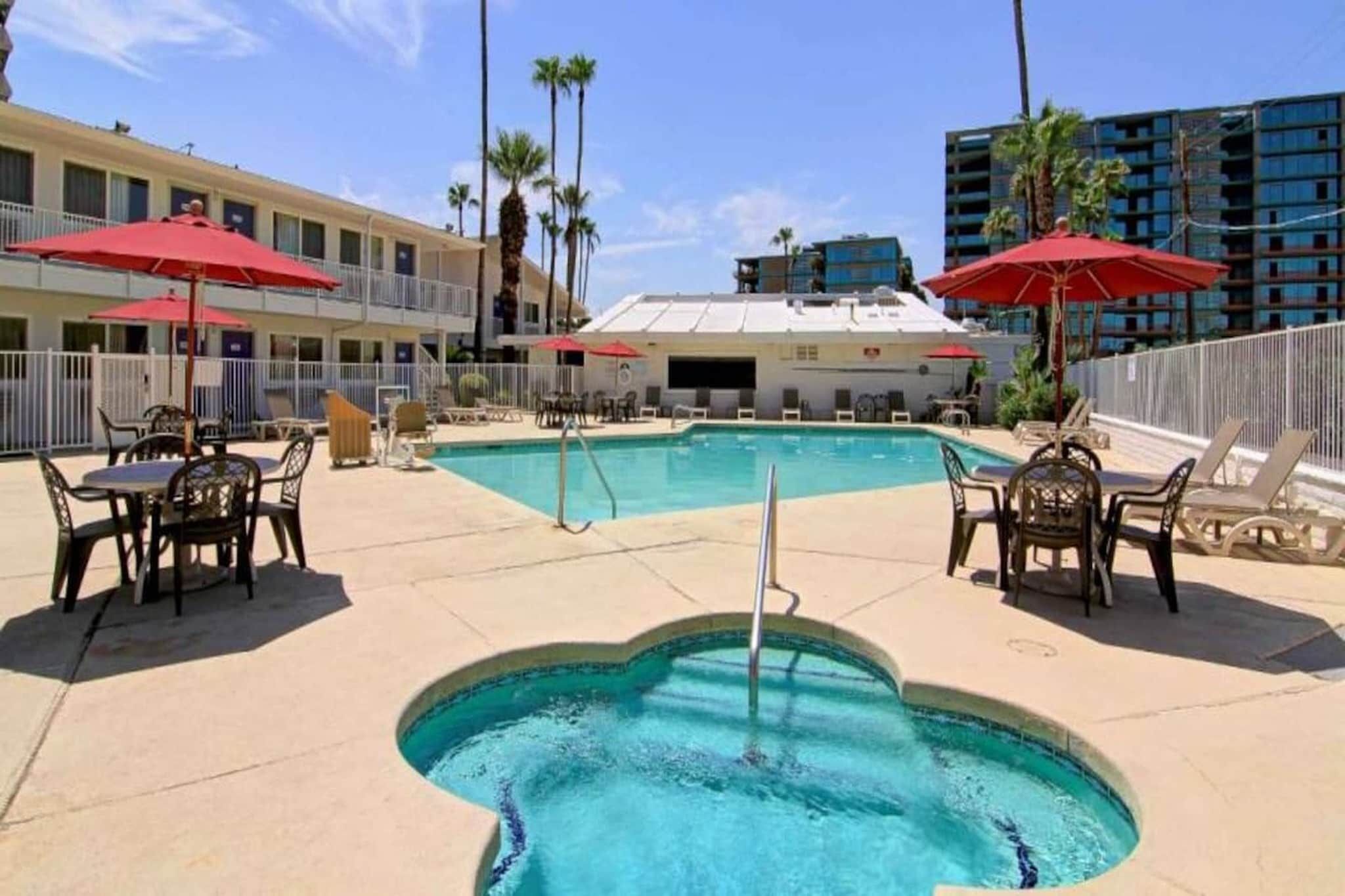 A swimming pool with lounge chairs and umbrellas, surrounded by buildings and palm trees.