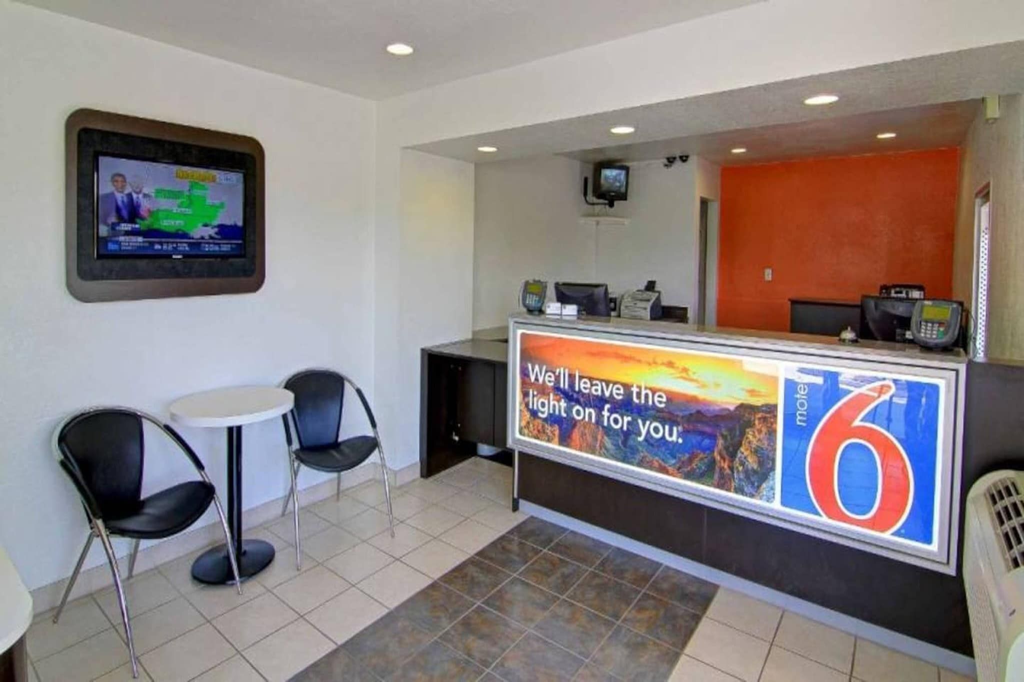 A hotel reception area with a counter, two chairs, a television screen, and a sign with a message.