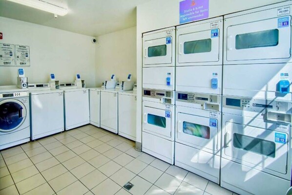A laundry facility with multiple washing machines and dryers, a tiled floor, and signs indicating service areas. - Studio 6 Phoenix, Az - Deer Valley_1 Kin (PHOENIX)