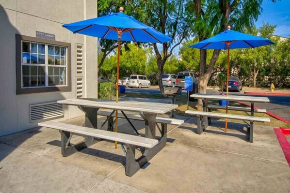 Mesas de picnic al aire libre con sombrillas azules, un edificio con cartel de lavandería y coches aparcados al fondo.