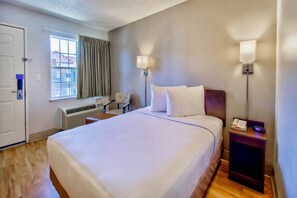 Una habitación de hotel con cama, mesita de noche, lámpara, ventana, equipo de aire acondicionado y una silla.