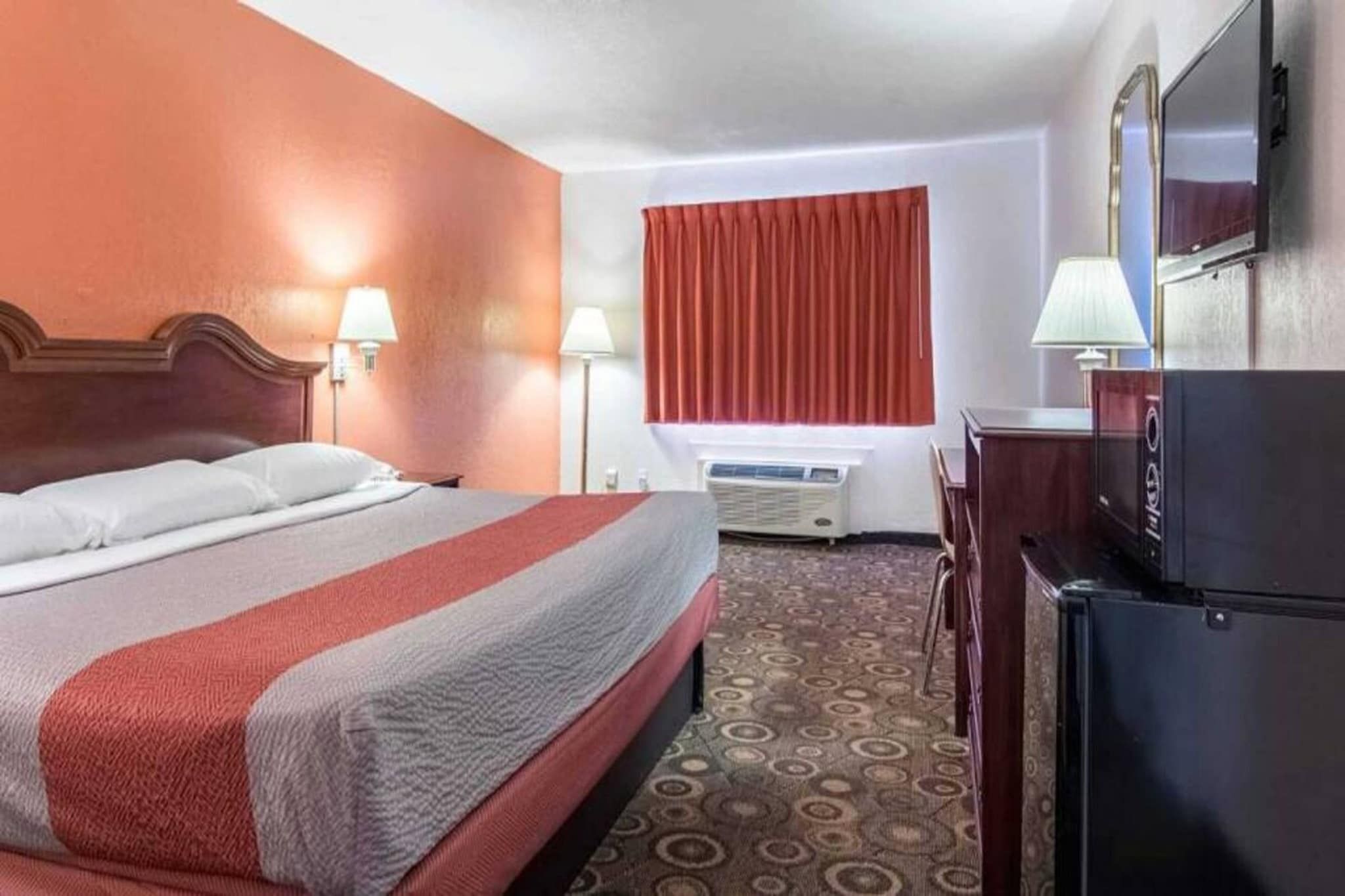 A hotel room with a bed, a desk, a refrigerator, a window with curtains, and an air conditioning unit.