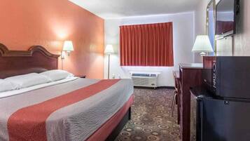 A hotel room with a bed, a desk, a refrigerator, a window with curtains, and an air conditioning unit.