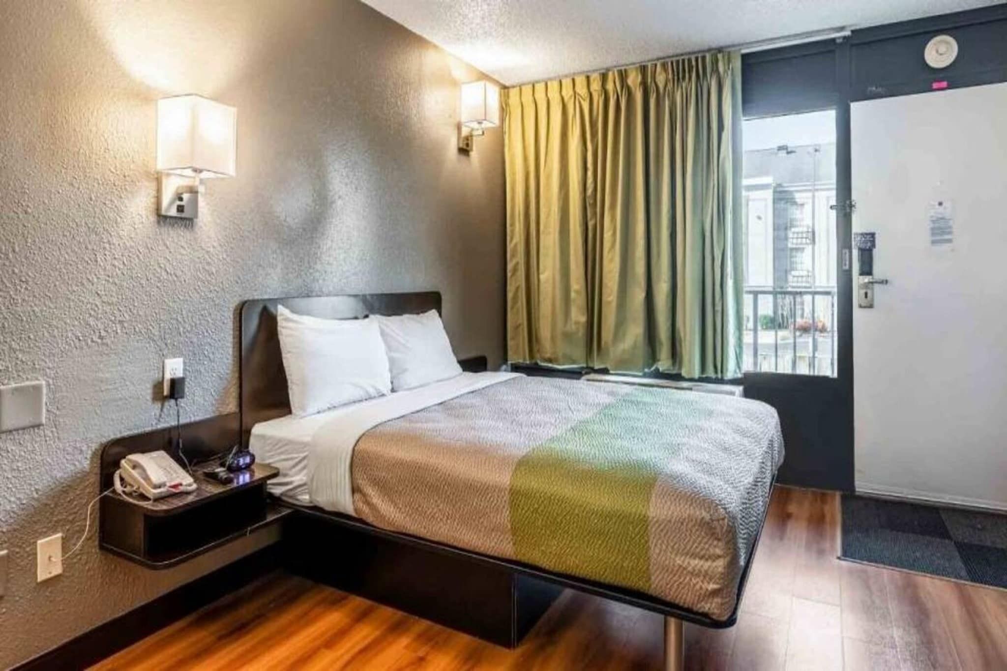 Une chambre d’hôtel avec un lit, une table de chevet, une lampe et une vue sur l’extérieur par une fenêtre avec des rideaux.