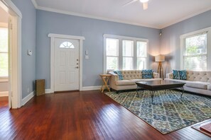 Une pièce avec un plancher en bois, une porte blanche, un canapé beige agrémenté de coussins à motifs, une table basse et un lampadaire.