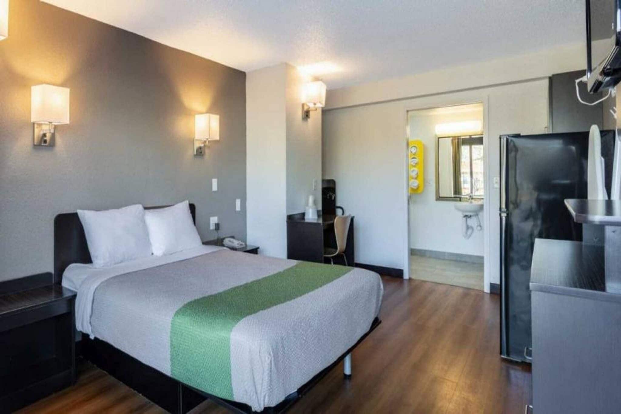 A hotel room with a bed, bedside tables, a desk, a chair, a refrigerator, and a bathroom with a sink and mirror.