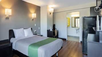 A hotel room with a bed, bedside tables, a desk, a chair, a refrigerator, and a bathroom with a sink and mirror.