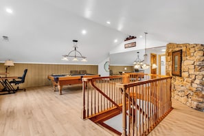 Una habitación amplia con mesa de billar, una escalera de madera y una pared de piedra.