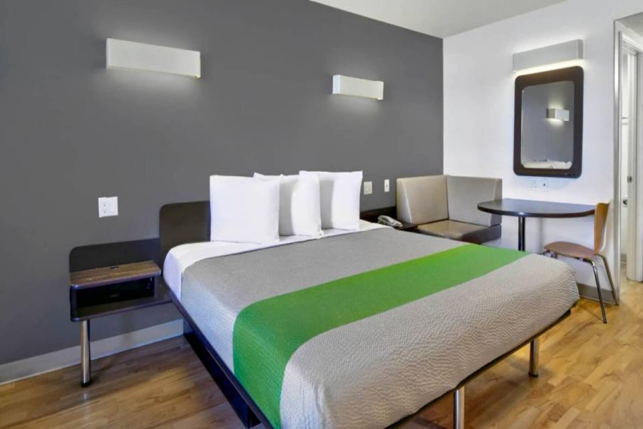 Quarto de hotel com cama, mesa de cabeceira, cadeira e pequena escrivaninha com cadeira.