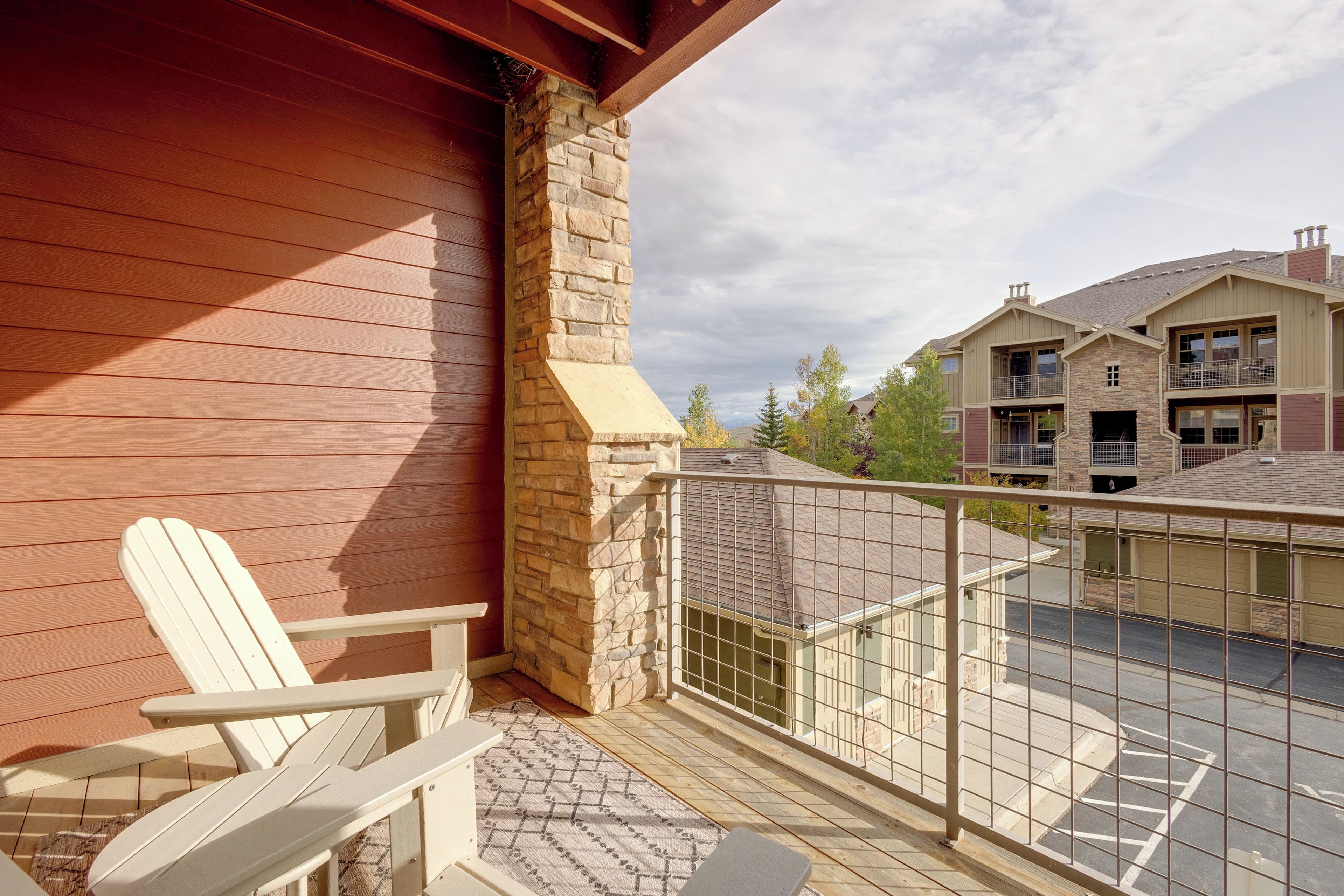A wooden deck with a white Adirondack chair, a stone chimney, and a view of neighboring buildings.