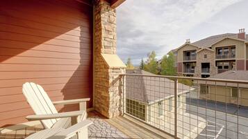 A wooden deck with a white Adirondack chair, a stone chimney, and a view of neighboring buildings.