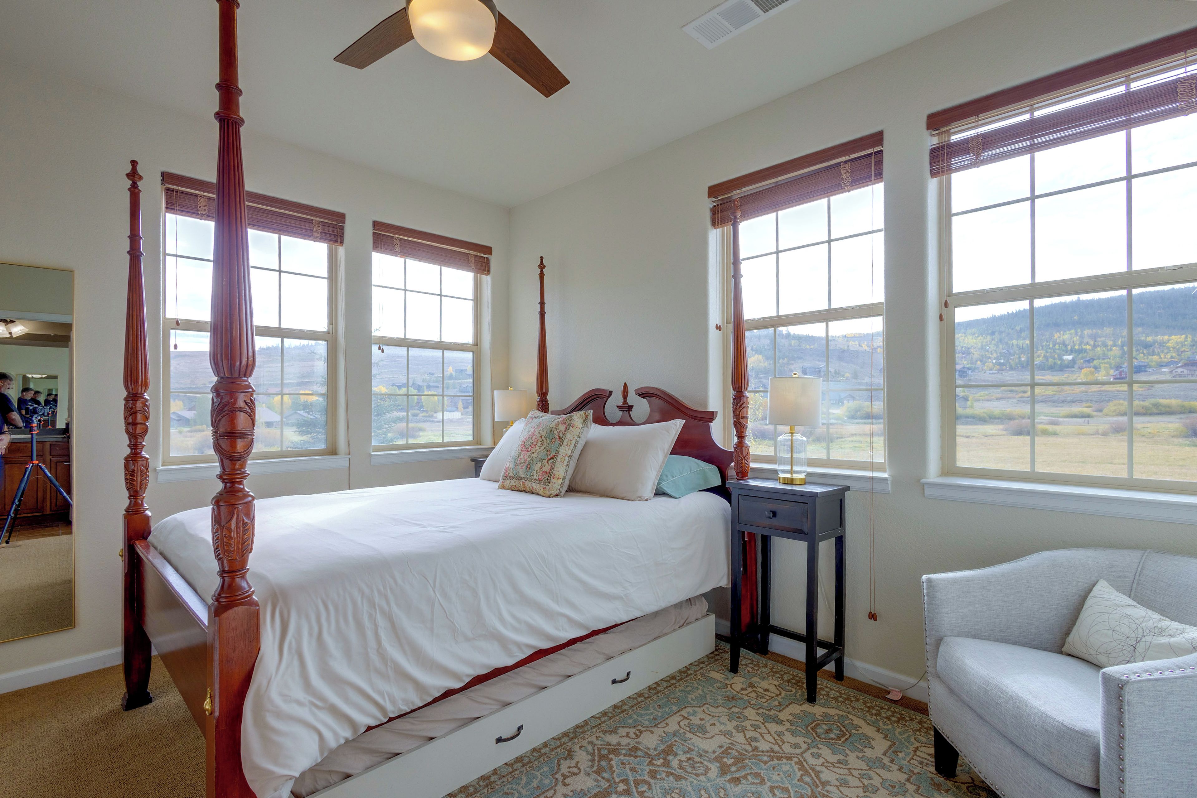 A four-poster bed with a wooden headboard, a bedside table with a lamp, a chair, and a view of the outdoors through large windows.