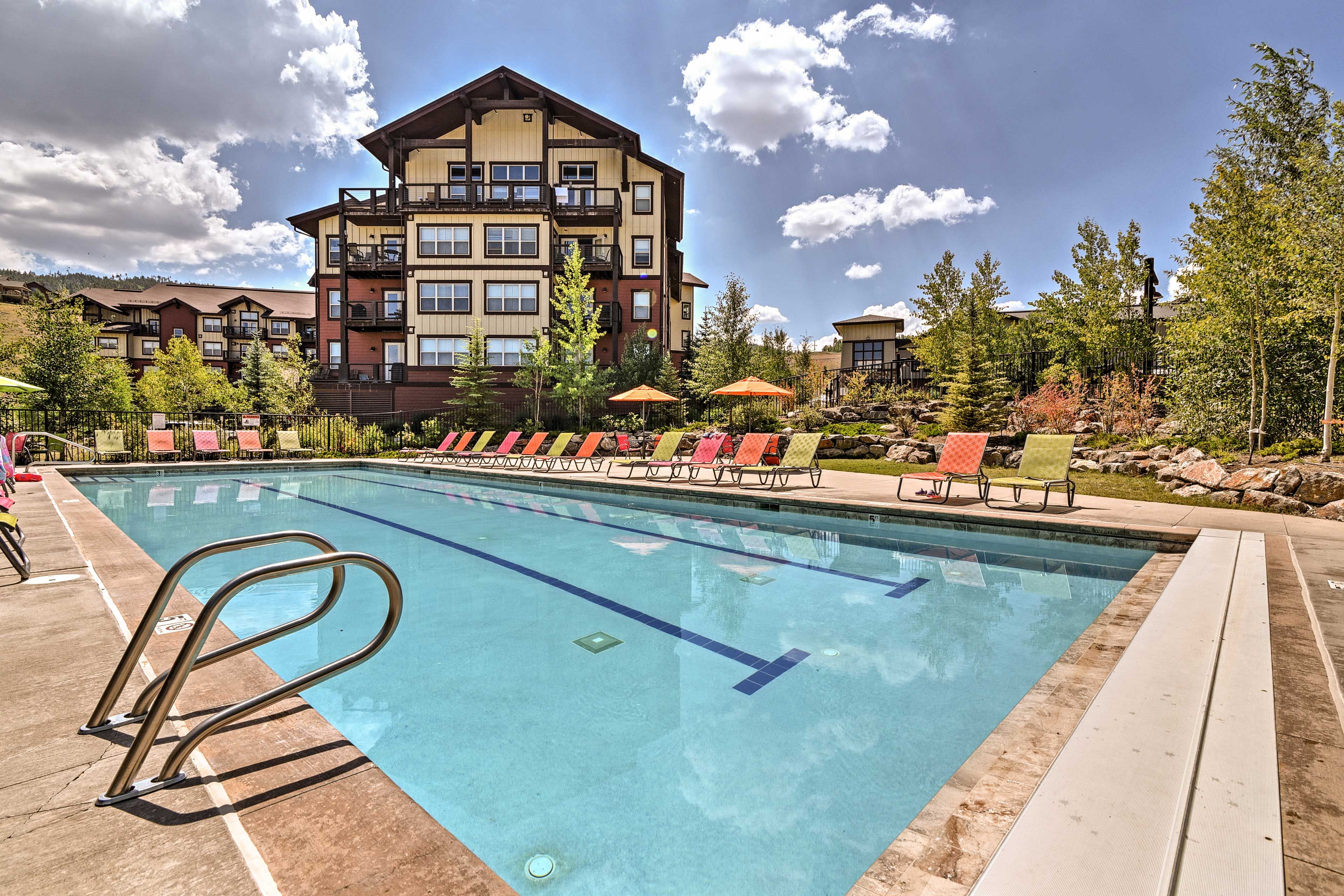 A swimming pool with a clear blue waterline, surrounded by lounge chairs and umbrellas, with a multi-story building in the background.