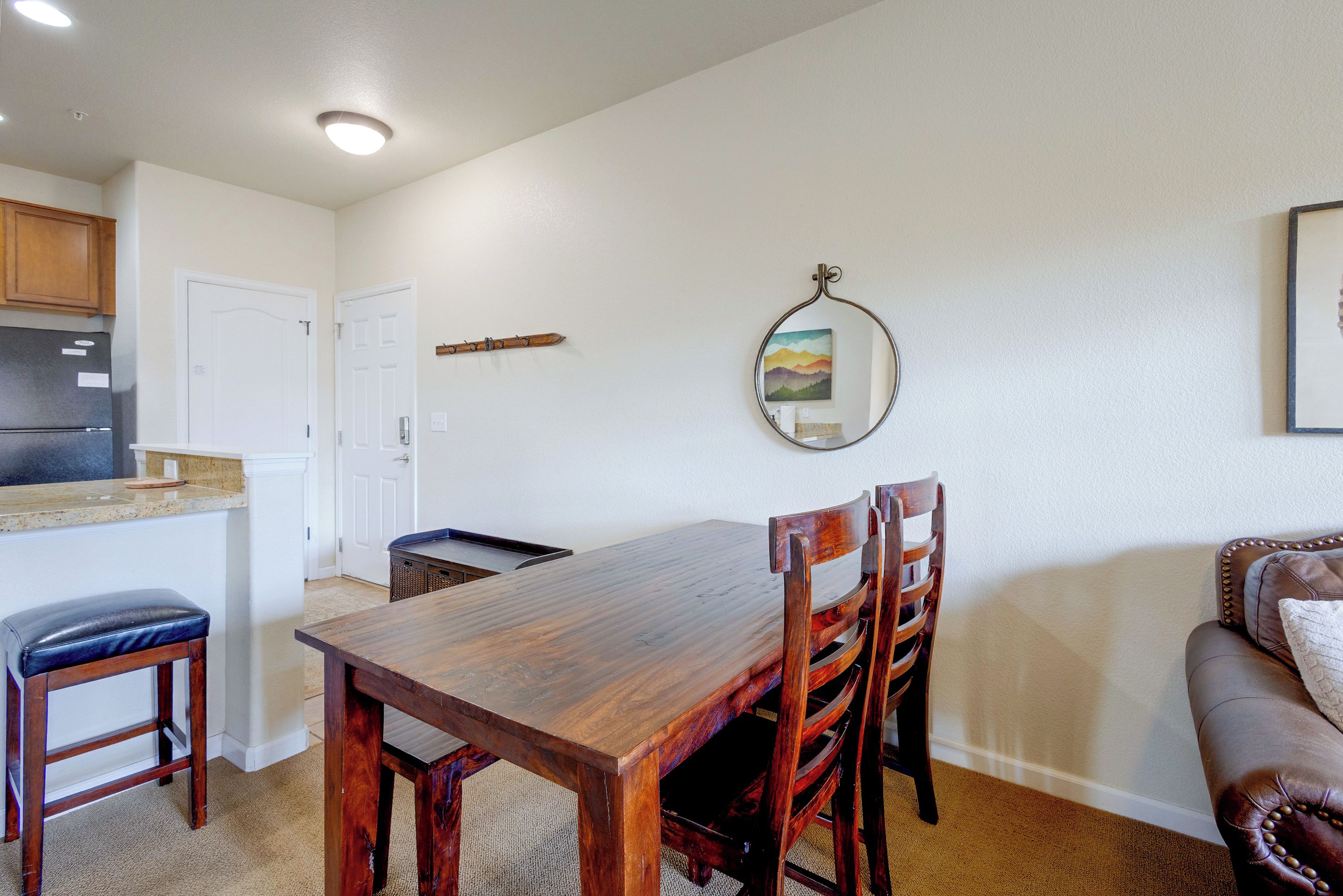 A dining area with a wooden table and chairs, a kitchen with a refrigerator, and a wall-mounted mirror.
