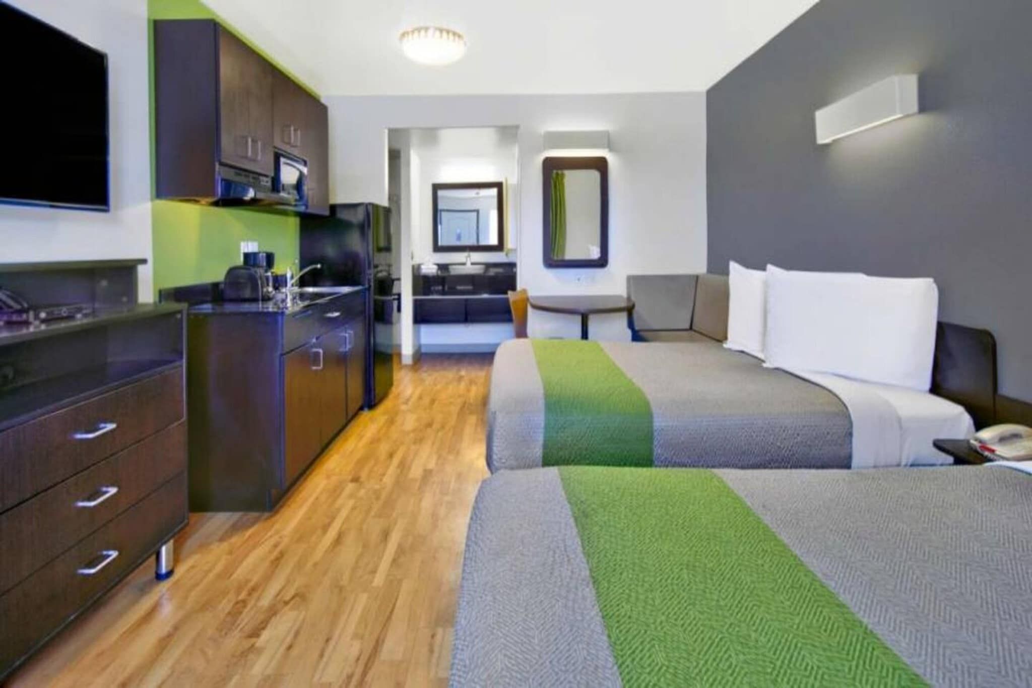 A hotel room with a kitchenette, a bed with two pillows, a bedside table, and a mirror.