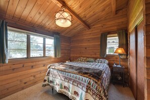 Quarto de cabana de madeira com uma cama, uma mesinha de cabeceira, um abajur e uma janela com cortinas.