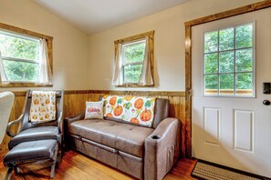 Una habitación acogedora con un sofá de cuero marrón, dos sillones y paneles de madera. Hay tres ventanas con cortinas, y una puerta blanca con picaporte.