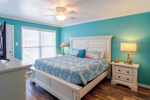 Une chambre à coucher comprenant un lit, des tables de chevet, une commode, un ventilateur de plafond et une fenêtre avec des stores.