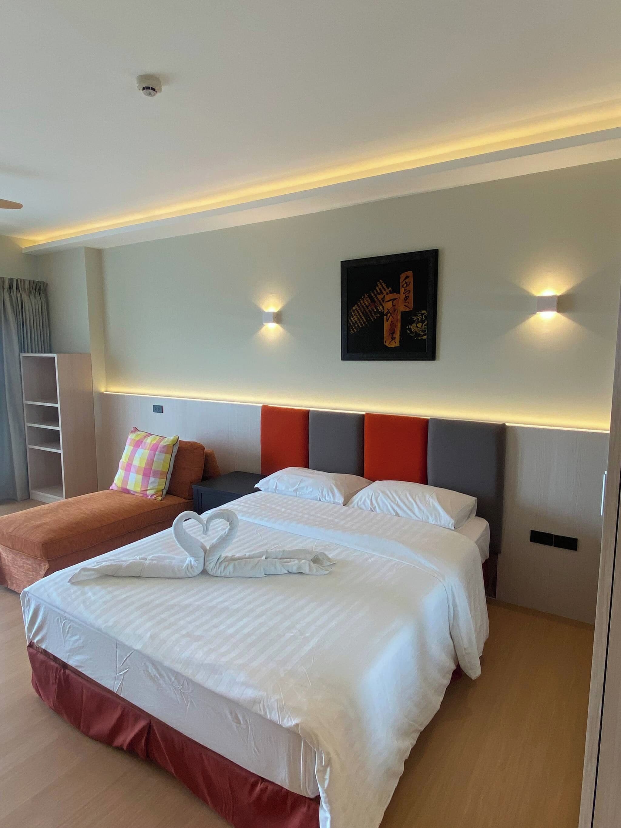 A hotel room with a large bed, a smaller sofa, a wall-mounted artwork, and a ceiling-mounted smoke detector.