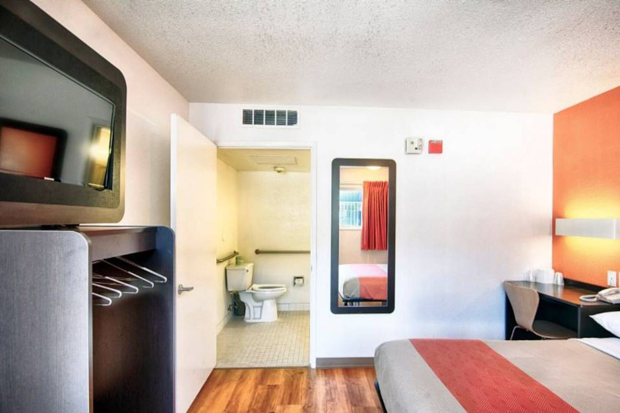 A hotel room with a bed, a desk, a chair, a mirror, and a bathroom visible through an open door.