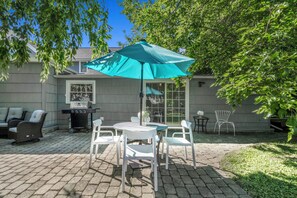 Een terras buiten met een barbecue, witte stoelen en een tafel onder een grote parasol.