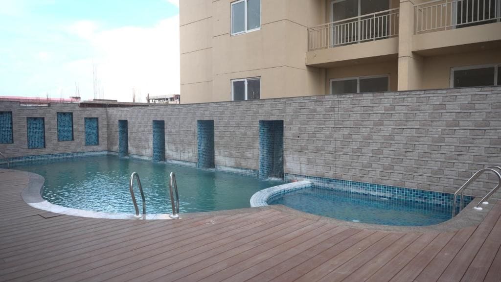 Uma piscina com deck de madeira, cercada por um muro de pedra, e adjacente a um edifício de vários andares.