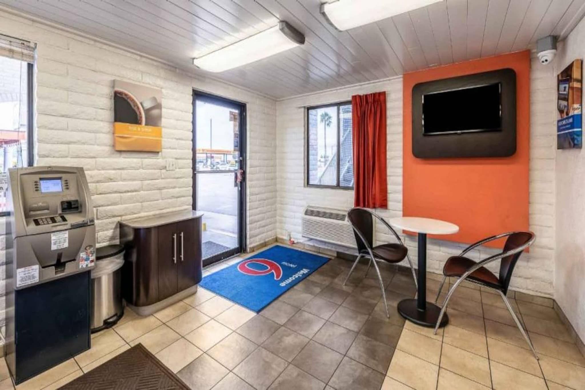 A small, modern room with a kitchenette, a seating area with a table and chairs, a television mounted on the wall, and a window with a view of the outside.
