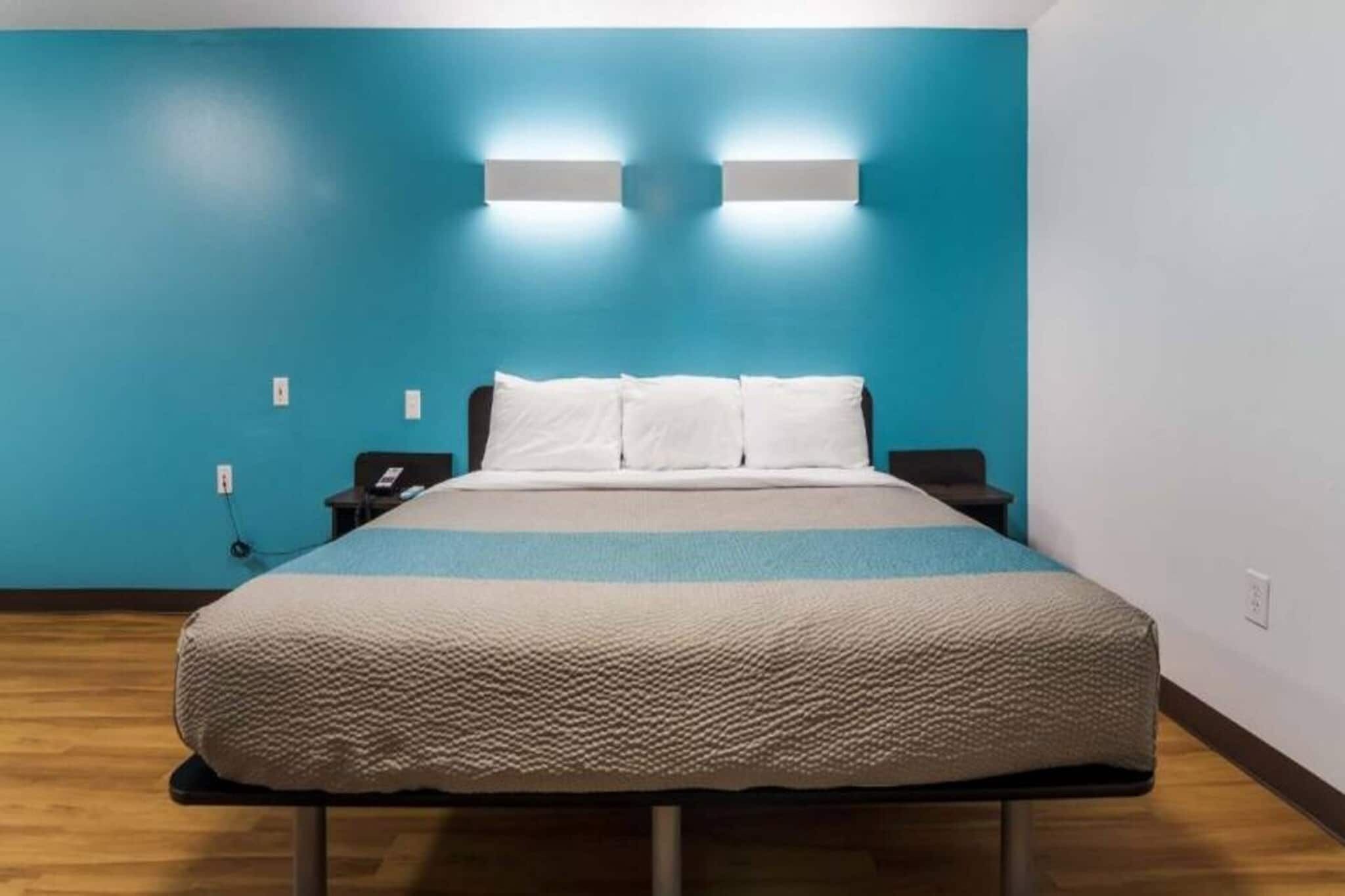 Een enkel bed in een kamer met een blauwe muur, een nachtkastje met een telefoon, en een wandlamp.
