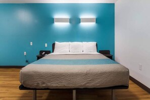 Een enkel bed in een kamer met een blauwe muur, een nachtkastje met een telefoon, en een wandlamp.