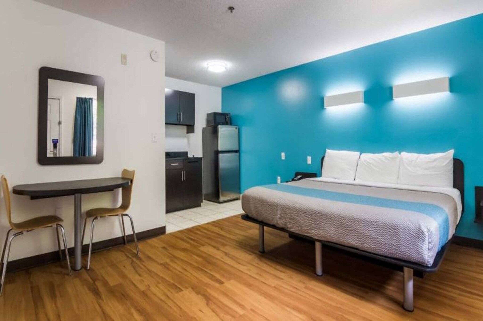 A hotel room with a bed, a desk, a chair, a mirror, a kitchenette, and a blue accent wall.