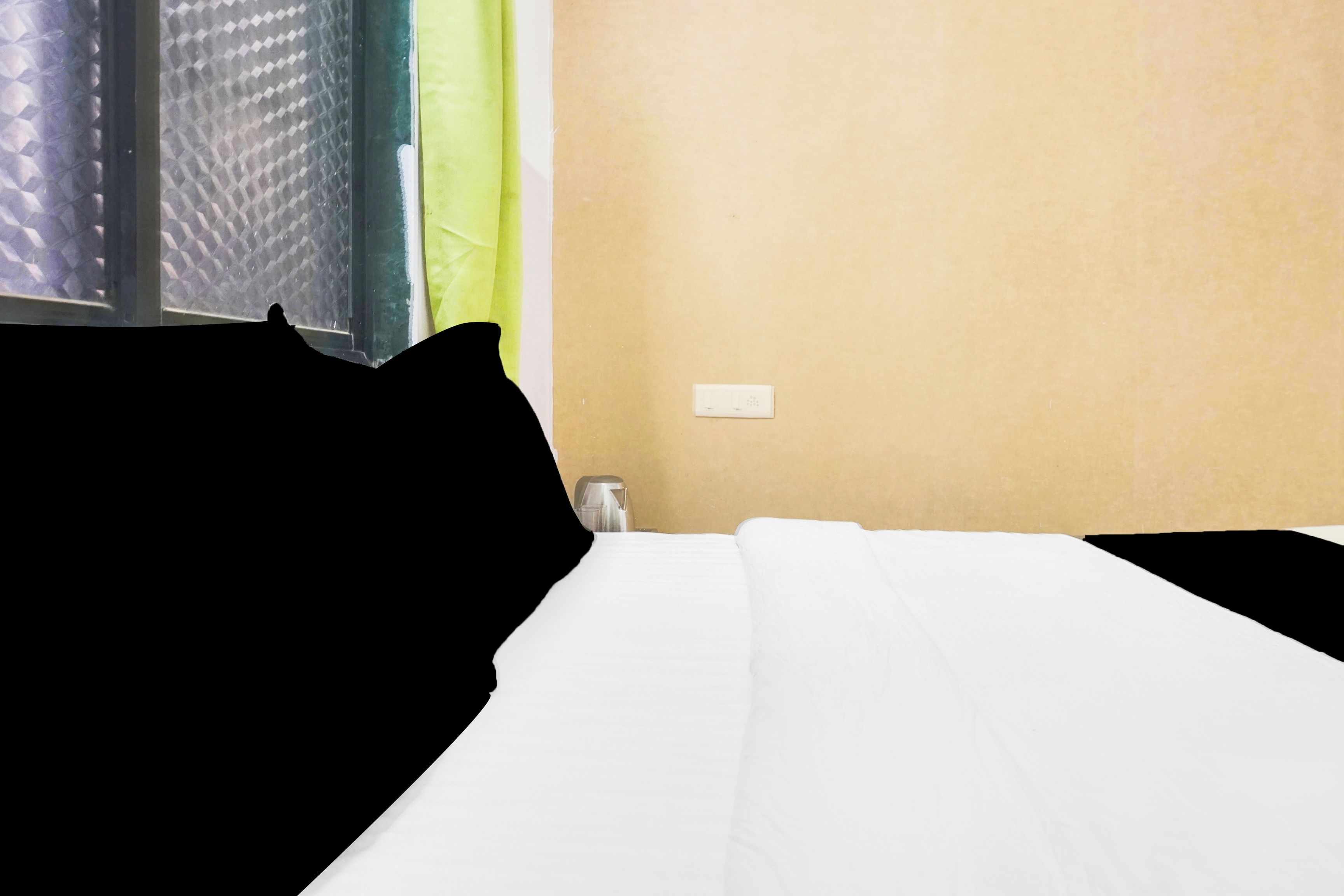Una habitación de hotel con una cama, un ventanal con persiana texturizada, un enchufe de pared y una pequeña mesa con una taza.