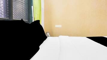 Una habitación de hotel con una cama, un ventanal con persiana texturizada, un enchufe de pared y una pequeña mesa con una taza.