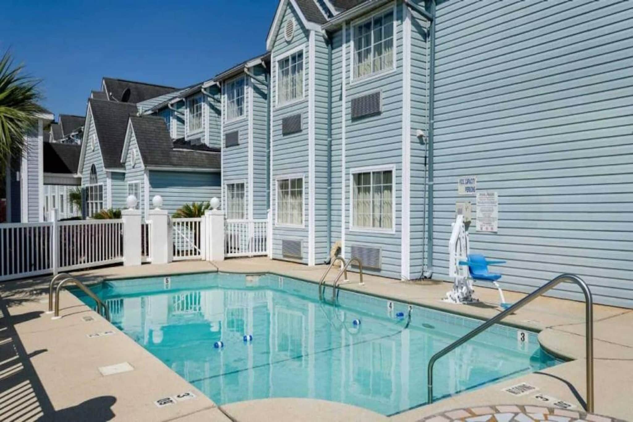 A swimming pool with a white fence, a blue building with white trim, and a clear sky.