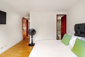 A hotel room with a bed, a standing fan, a wooden floor, a white wall, and a door leading to a bathroom. - Charming 2-Bedroom Apartment in Medellín's Golden Mile (Medellín)