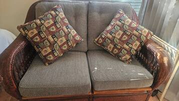 A wicker sofa with patterned cushions, positioned in front of a window with blinds.