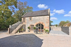 A stone building with a staircase leading to a door, a patio with outdoor seating, and a gravel driveway. - Butterton Barn (Leek)