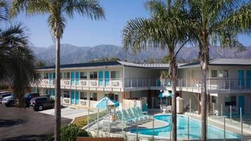 Hotel com piscina, palmeiras e montanhas ao fundo.