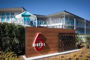 A modern motel with a prominent '6' sign, a covered outdoor seating area, and a balcony.