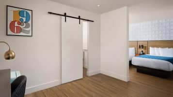 Quarto de hotel com cama, escrivaninha e cadeira. Possui porta deslizante e luminária fixada na parede.