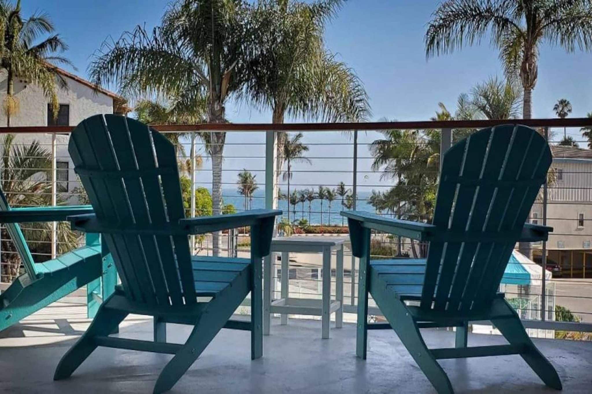 Two Adirondack chairs facing each other with a view of palm trees and a building in the background.
