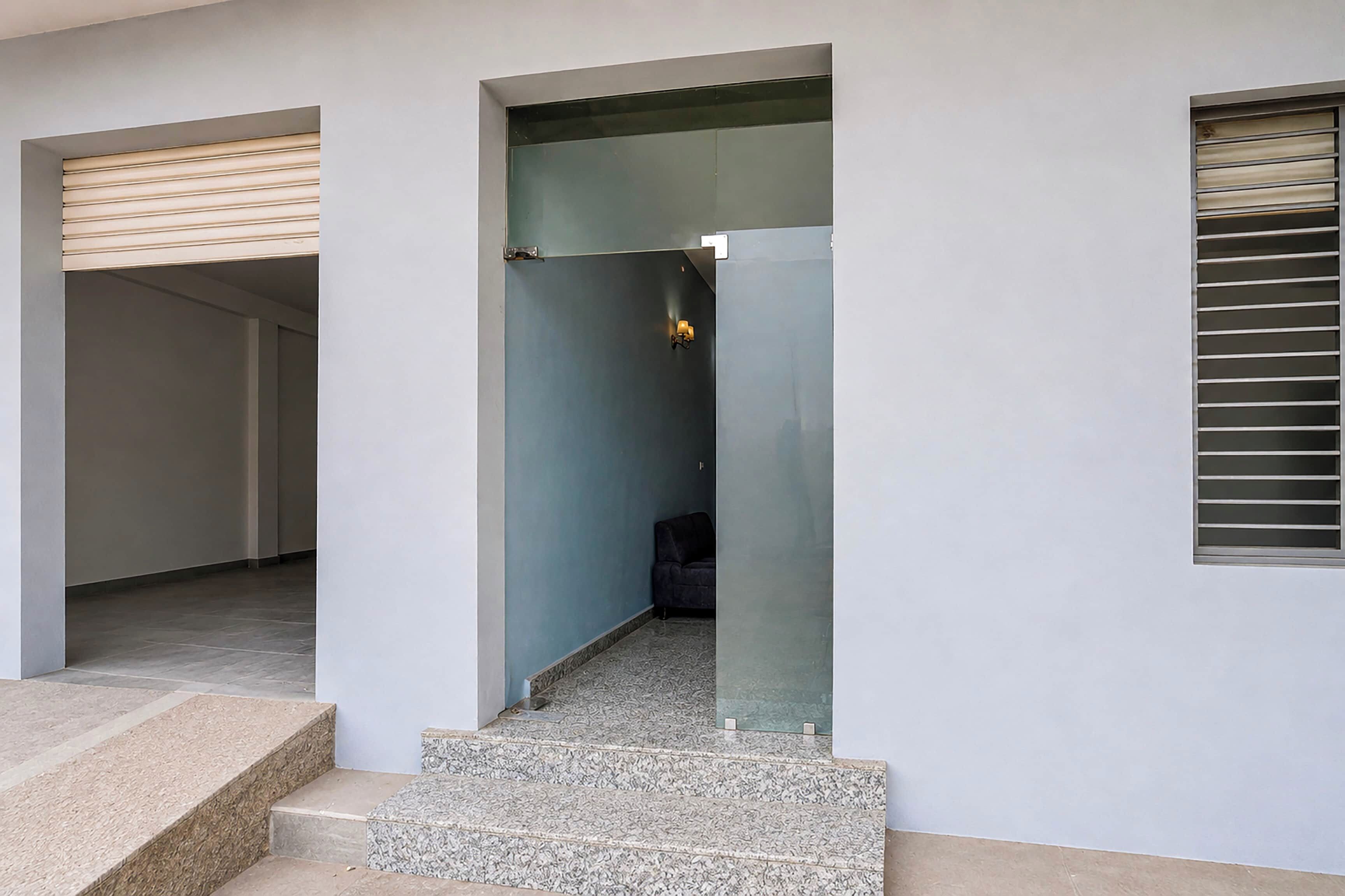 Entrada de un edificio moderno con puerta de vidrio, unos pocos escalones y un sofá de color oscuro en el interior.