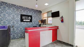 Zona de recepción de hotel con un mostrador rojo y blanco, un cartel con el nombre del hotel, un extintor de incendios en la pared y un papel tapiz decorativo.