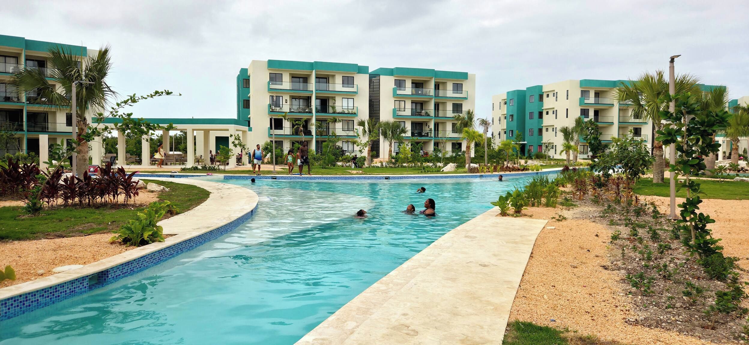 Una piscina con gente nadando, rodeada por un edificio con balcones y palmeras.