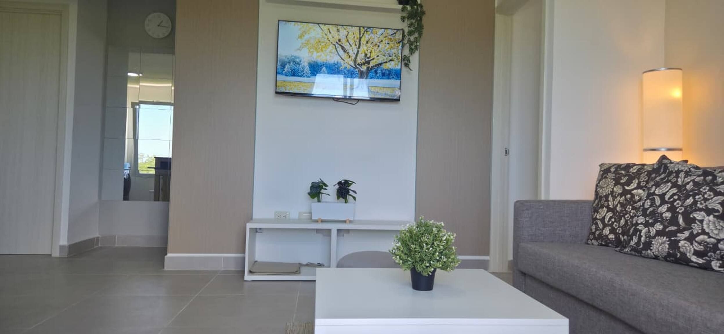 Una sala de estar con una mesa de centro blanca, un sofá gris con un cojín estampado, un cuadro de un árbol y un reloj de pared.