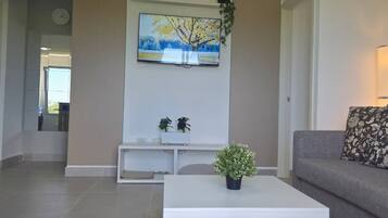 Una sala de estar con una mesa de centro blanca, un sofá gris con un cojín estampado, un cuadro de un árbol y un reloj de pared.