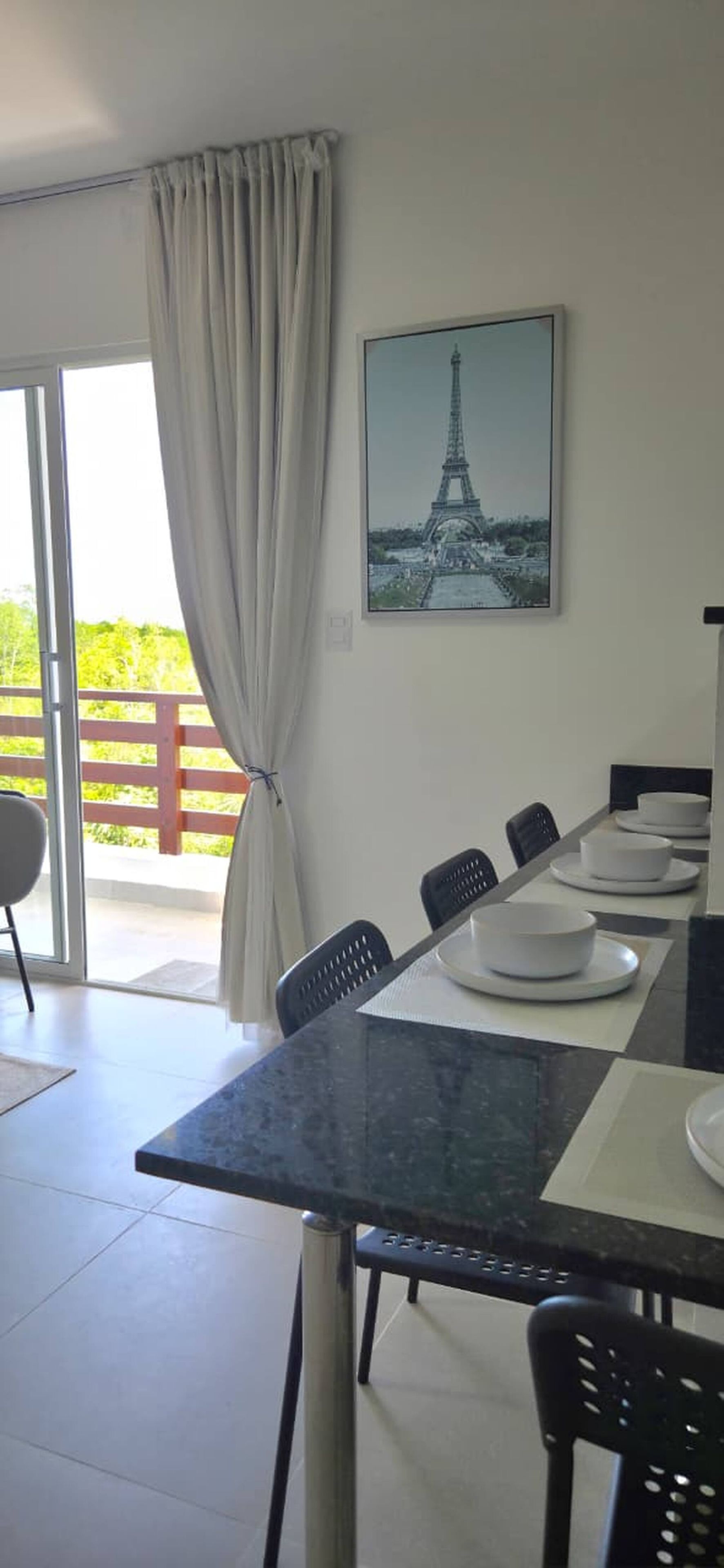 Comedor con una mesa dispuesta para cuatro, sillas, un cuadro de la Torre Eiffel y vistas a una zona exterior con terraza de madera.