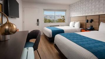 A hotel room with two beds, a desk, a chair, a TV, and a window with a mountain view.