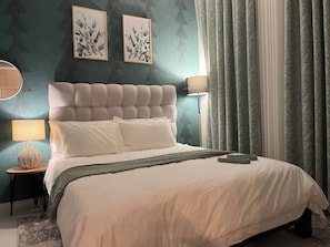Une chambre à coucher avec une tête de lit capitonnée, un lit recouvert de draps blancs, deux lampes fixées au mur et une grande fenêtre avec des rideaux.
